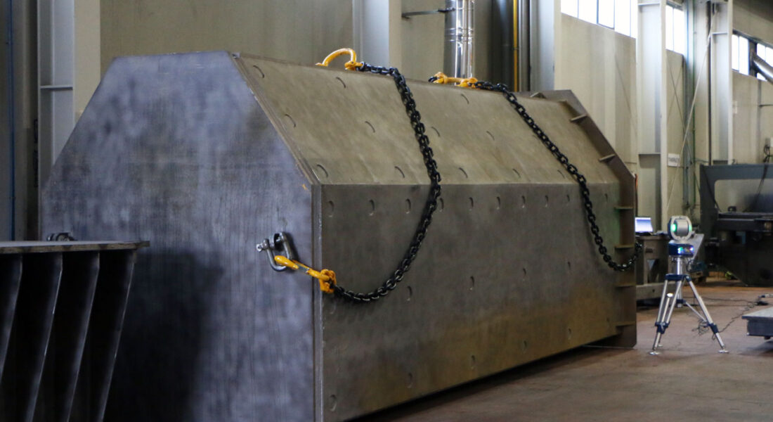 Gran soporte mecanizado posado en el suelo de las instalaciones de LAMUCE y con cadenas de transporte para puente grúa de 40t. Vista lateral izquierda del soporte. Detalle de la pieza con láser faro en segundo plano